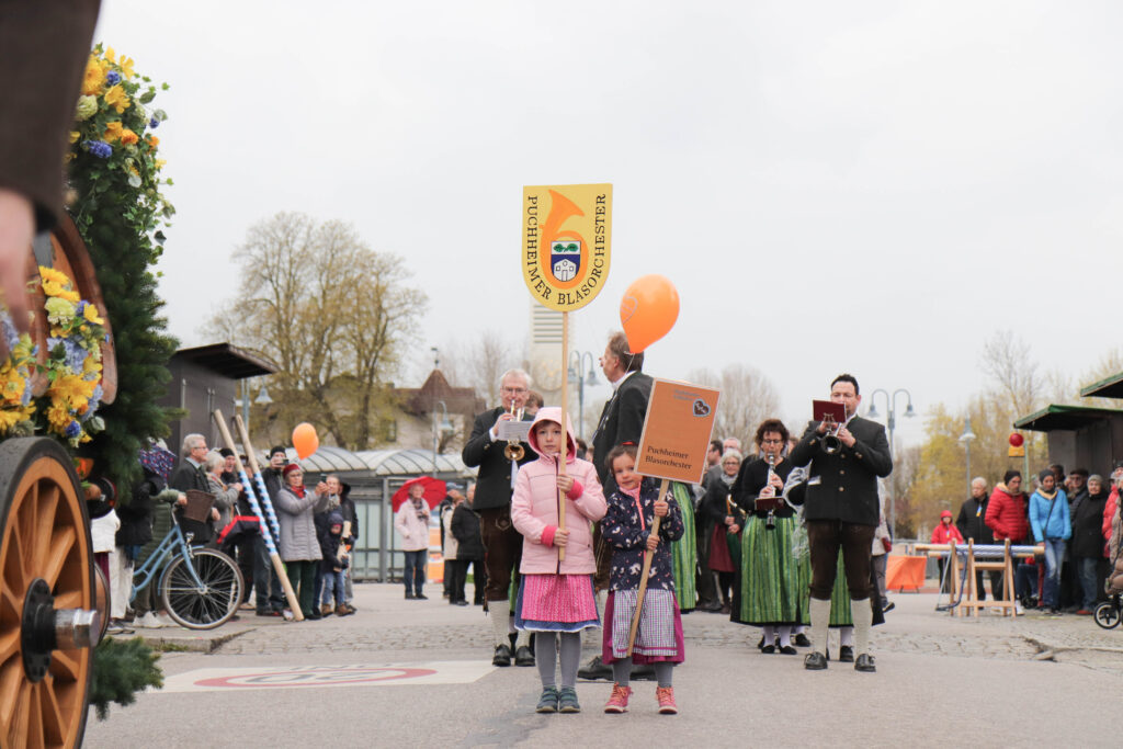 Die kleinen Besucher halten stolz das Vereinsschild des Puchheimer Blasorchesters, links und rechts stehen Besucher die die Musik des Blasorchesters genießen